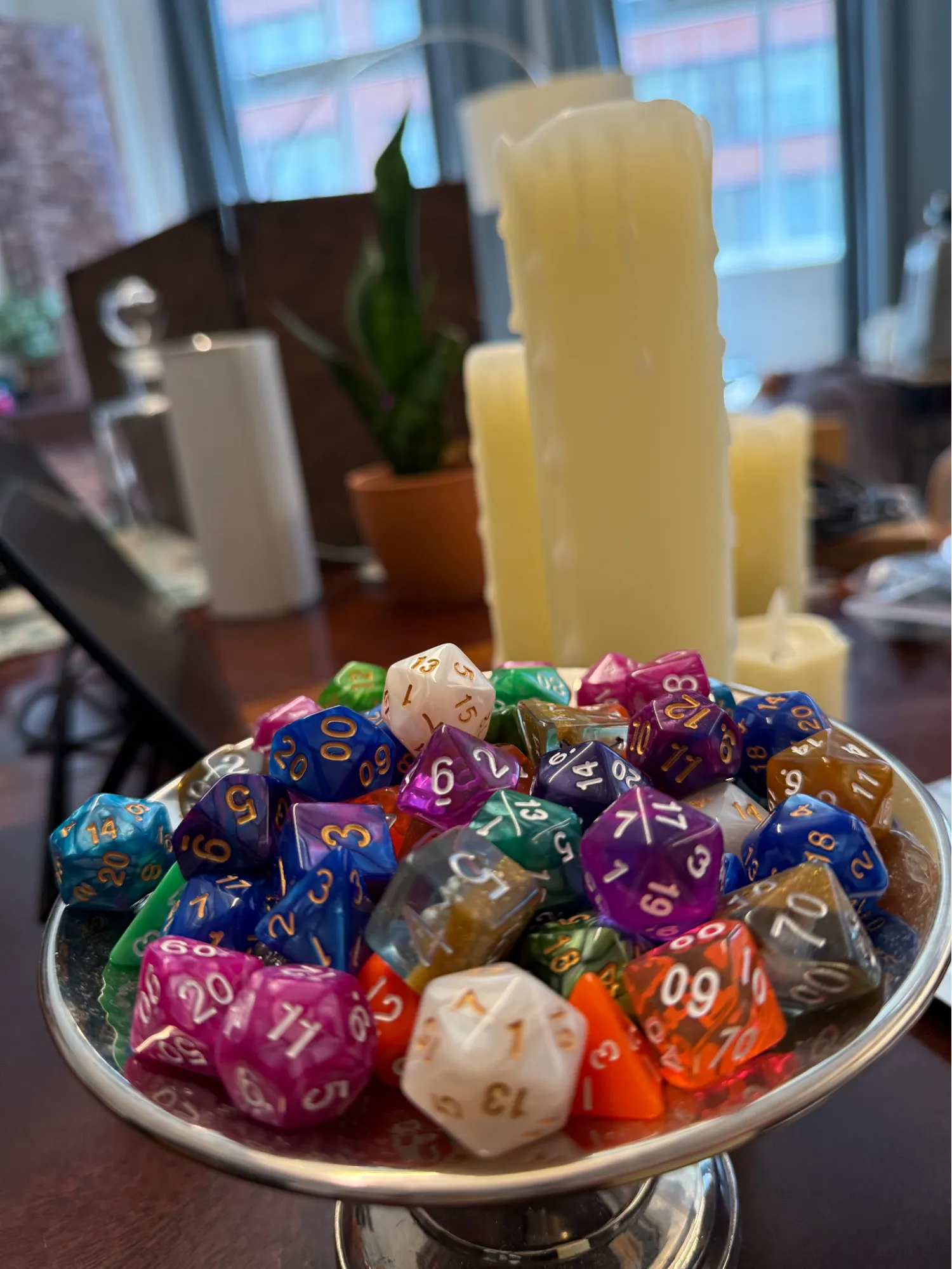 A silver pedestal dish piled with colorful polyhedral dice beside tall dripping candles